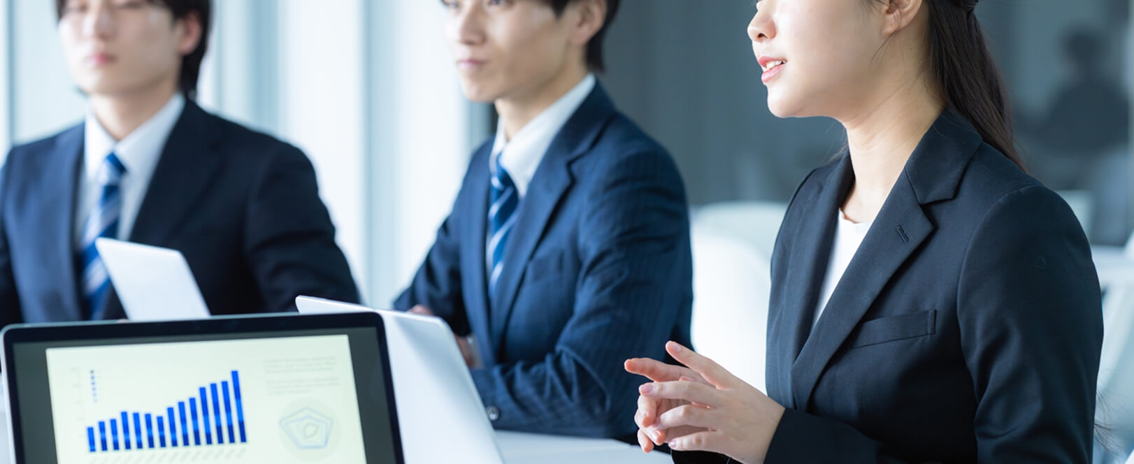 会議室でグラフ資料を見ながら発言者の話を聞いているスーツ姿の男女