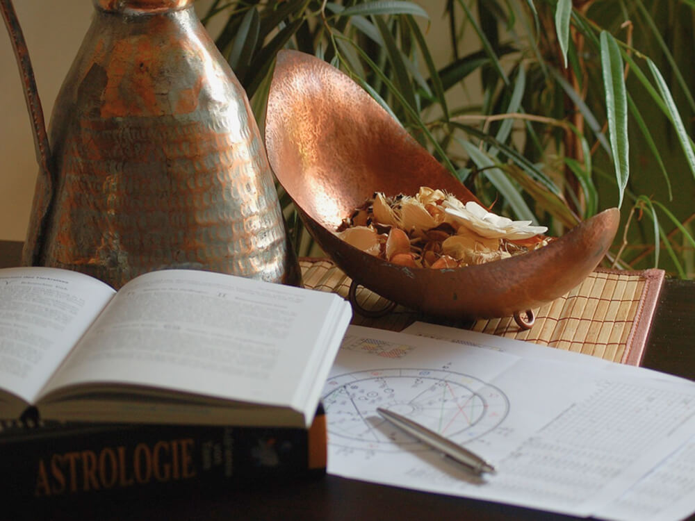 占星術の書籍とホロスコープの資料、銅製のポットとドライフラワーが置かれた落ち着いた机の上の風景